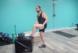 Dr. John standing with his left foot elevated on a box. He is holding a bow muscle scraper in his hands and is applying pressure to his hamstrings with it as a form of pulled hamstring treatment